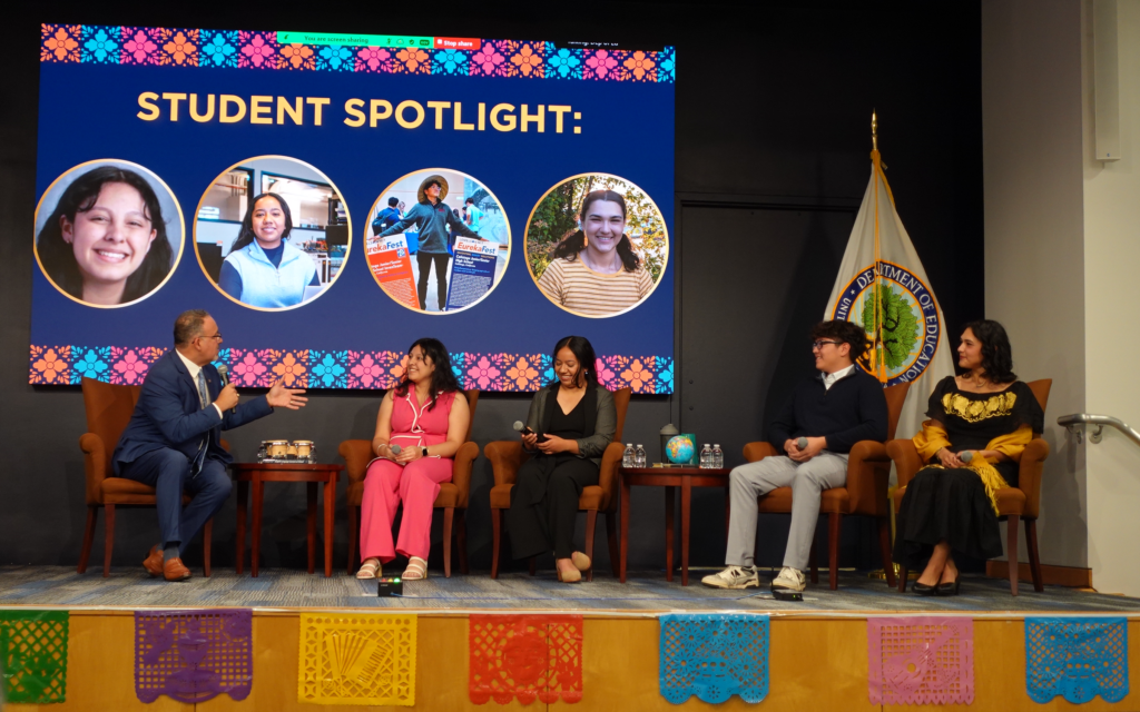 U.S. Secretary of Education Miguel Cardona is sitting on the right of the stage holding a microphone. He is leading a panel discussion with four student inventors seated nearby. Next to him is a young female inventor, then LMIT InvenTeam alum Lesly Rojas, and Calistoga InvenTeam member Elias Escobar, and InvenTeam alum Katia Avila. At the front of the stage floor, there are colorful flags, and behind the panelists is a big screen that reads “Student Spotlight” and shows pictures of the panelists. The U.S. Department of Education flag is also on display behind them.
