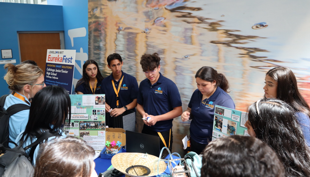 Five students from the Calistoga InvenTeam are standing behind a table, showing their invention project to other students at EurekaFest. There are posters and other materials related to their project on the table in front of them.
