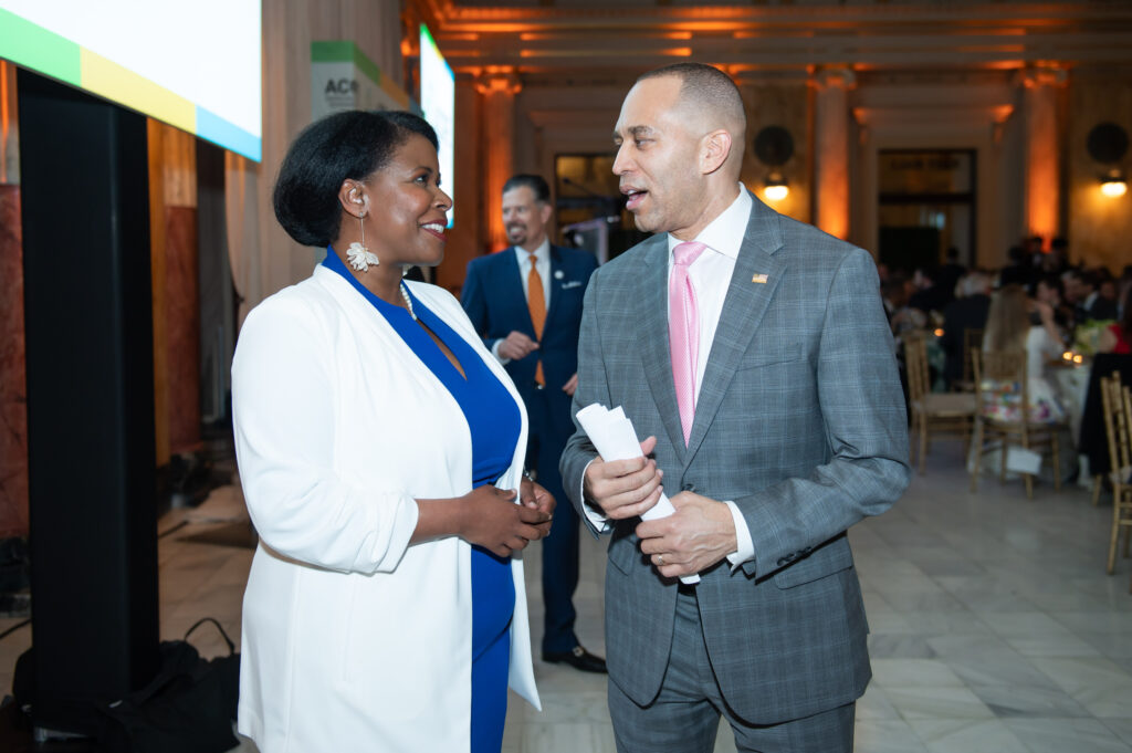 A woman in a white blazer speaks with a congressman in a gray blazer at an event.