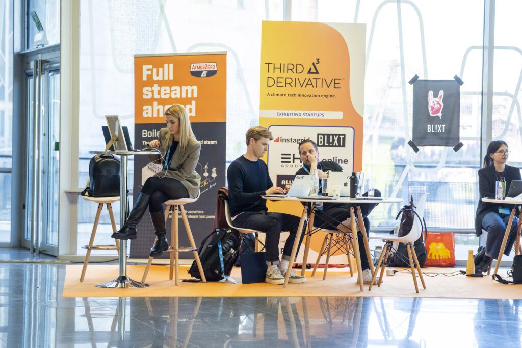 Individuals working at laptops in front of orange signs that say, "Third Derivative," a portfolio company of Rocky Mountain Institute at the Energy Tech Summit.